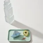 Flat-lay still life: frosted glass tower looming over a small pastel-blue paper sailboat on a mint tray, symbolizing enterprise ownership over an indie tool