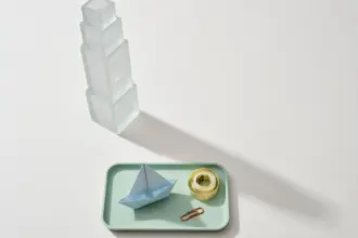 Flat-lay still life: frosted glass tower looming over a small pastel-blue paper sailboat on a mint tray, symbolizing enterprise ownership over an indie tool
