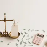 Flat-lay: vintage brass balance scale with gold coins on one pan and a paper invoice tag on the other, surrounded by pastel office props