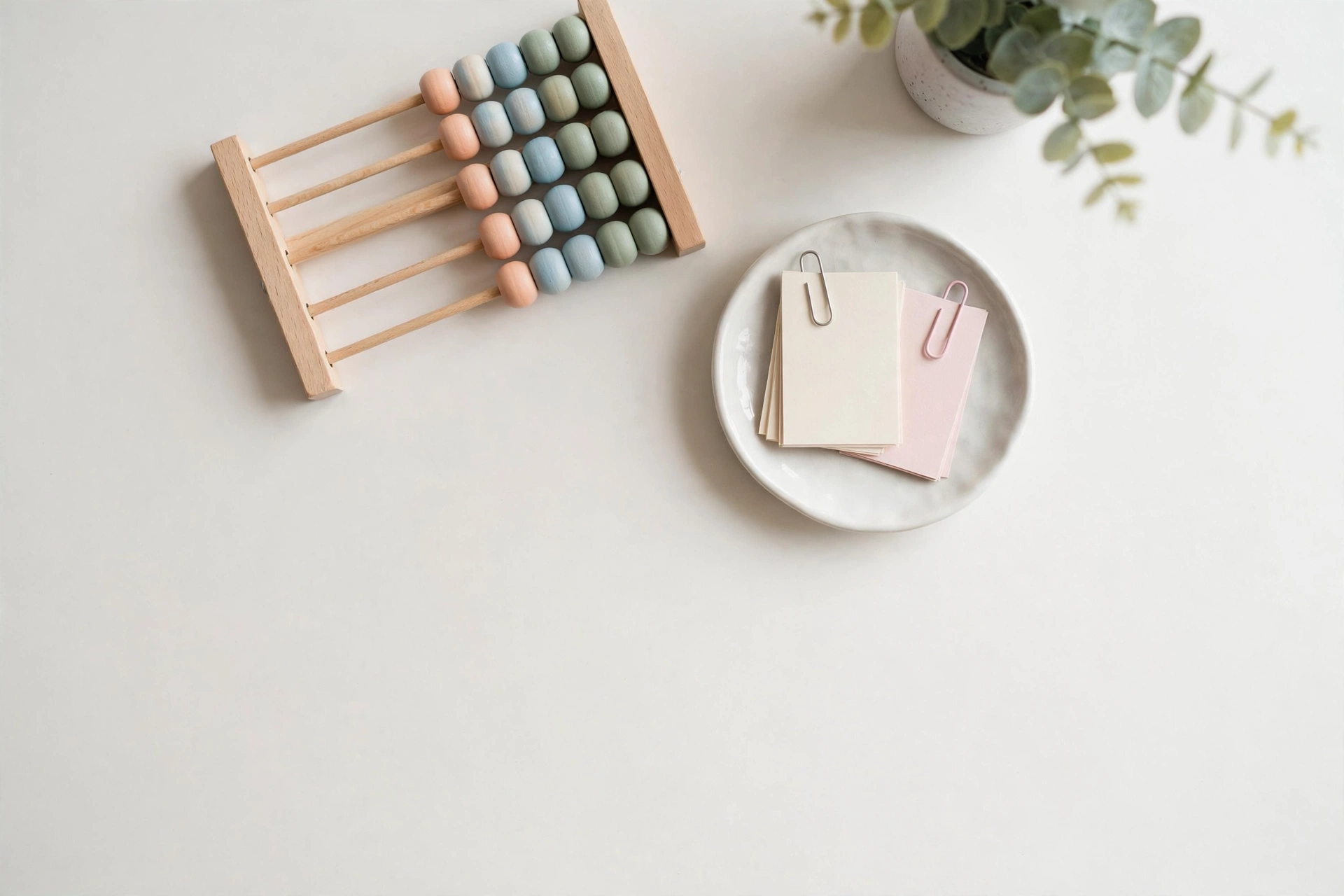 Flat-lay: miniature wooden abacus with pastel beads beside index cards and a eucalyptus sprig, symbolizing fee calculations