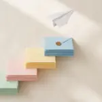 Flat-lay: four-step staircase built from stacked pastel envelopes ascending left to right with a brass coin on the top step and a paper airplane above