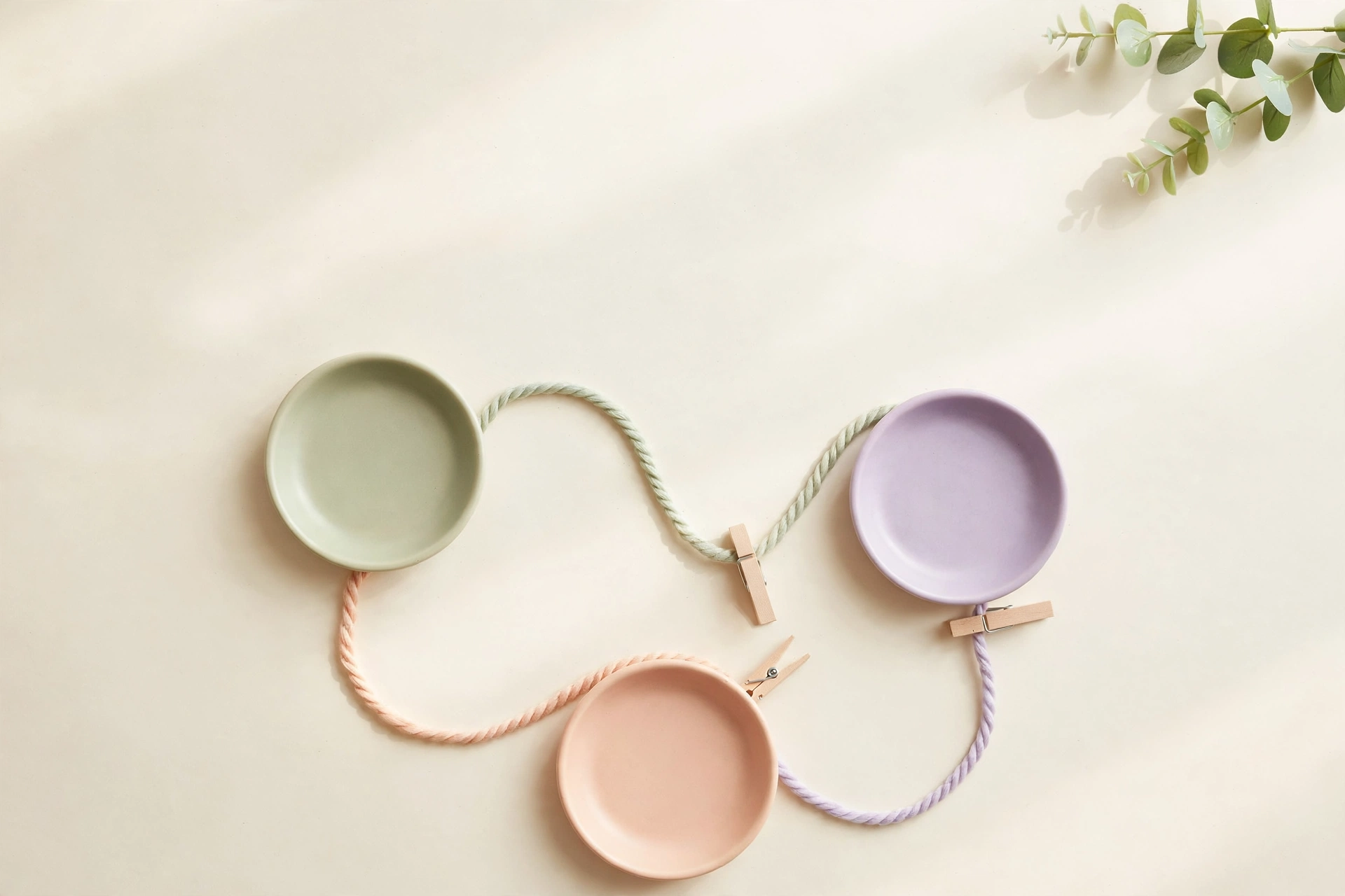 Flat-lay: pastel switchboard of three ceramic dishes connected by looping yarn threads with wooden clothespins, symbolizing automation wiring