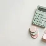 Flat-lay: small calculator beside three stacked pastel coins and a tiny paper invoice, mint green and blush pink palette