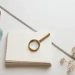 Flat-lay: brass magnifying glass on a stack of cream paper sheets with a chain of silver links running diagonally, symbolizing information linked into a chain