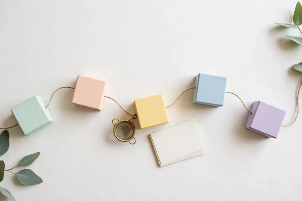 Flat-lay: five pastel wooden blocks in an arc connected by twine thread with a tiny brass sieve beside, symbolizing pipeline stages with a verification filter