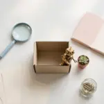 Flat-lay: open kraft box with a brass robot figurine peeking out beside a tiny succulent, surrounded by pastel research props