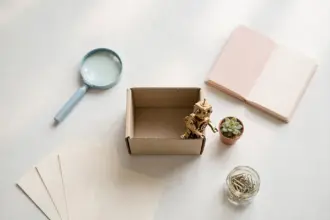 Flat-lay: open kraft box with a brass robot figurine peeking out beside a tiny succulent, surrounded by pastel research props