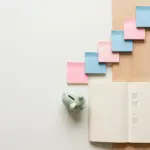 Flat-lay: small wooden desk scene with a cream notebook open to a checklist, a mint-green piggy bank, and pastel sticky notes in a rising staircase