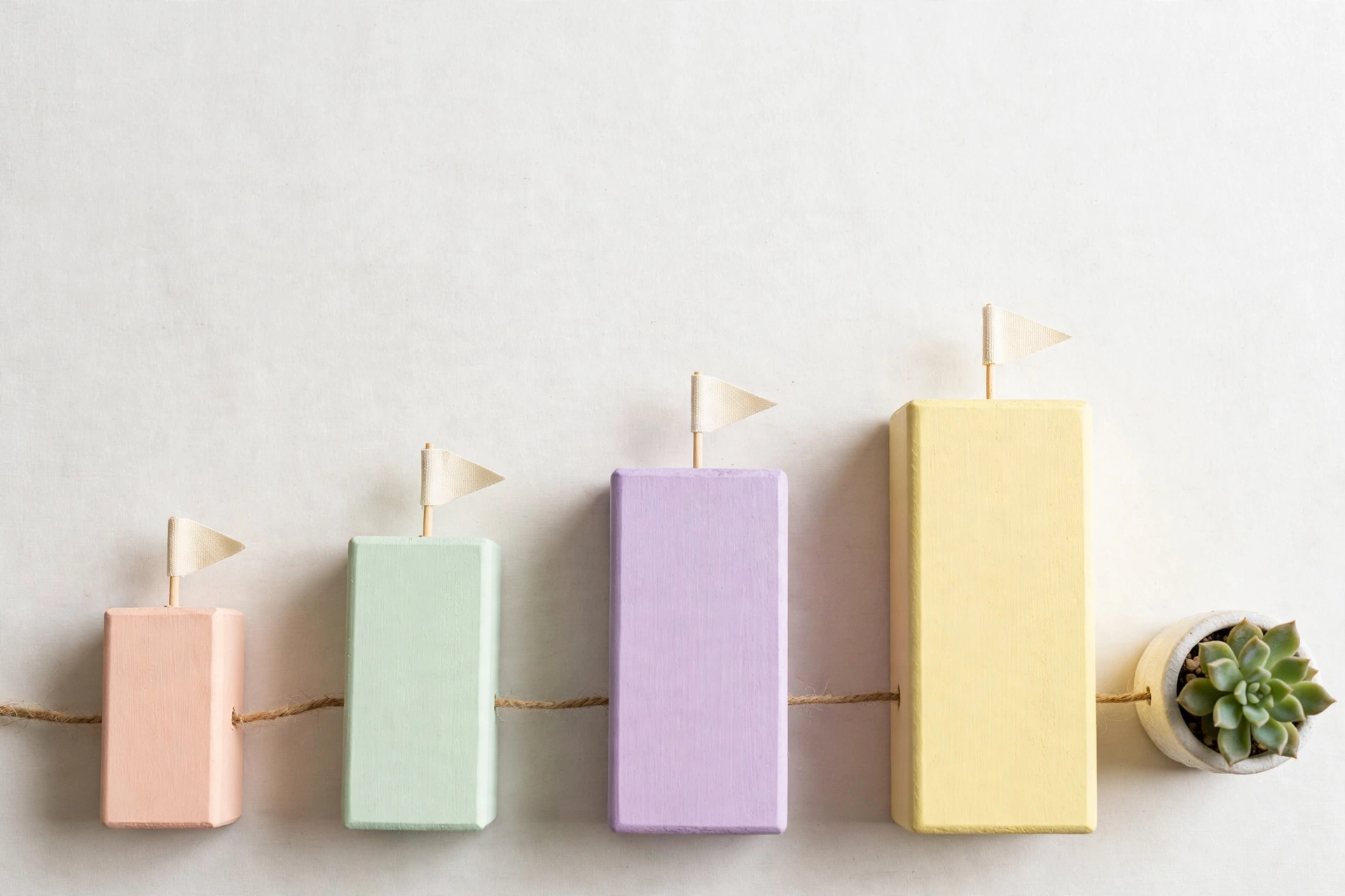 Flat-lay: neat row of pastel wooden building blocks in ascending sizes, each topped with a tiny cream flag, connected by twine