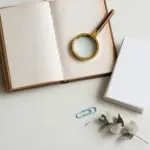 Flat-lay: open vintage hardcover book with a small magnifying glass resting on the page, a stack of white index cards beside it