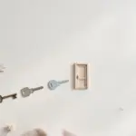 Flat-lay: three different keys — a vintage brass key, a grey key and a pale blue key — in a row leading to a tiny wooden door on a pale off-white background, with a small pink dish and eucalyptus leaves at the edges