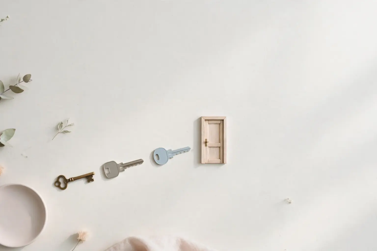 Flat-lay: three different keys — a vintage brass key, a grey key and a pale blue key — in a row leading to a tiny wooden door on a pale off-white background, with a small pink dish and eucalyptus leaves at the edges