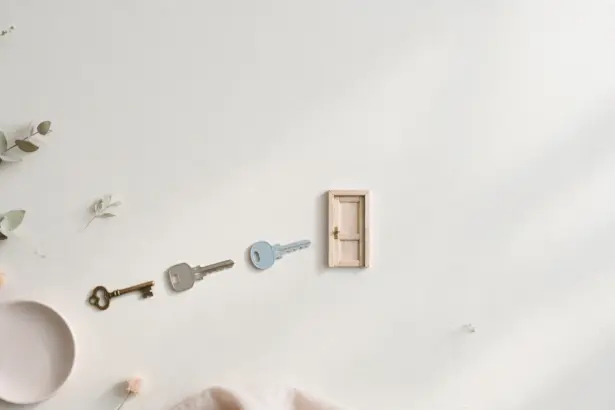 Flat-lay: three different keys — a vintage brass key, a grey key and a pale blue key — in a row leading to a tiny wooden door on a pale off-white background, with a small pink dish and eucalyptus leaves at the edges