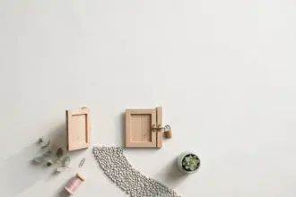 Flat-lay: two small wooden garden gates on a pale off-white background, one open and one closed with a tiny brass padlock, a curved pebble path between them and a small potted succulent beside