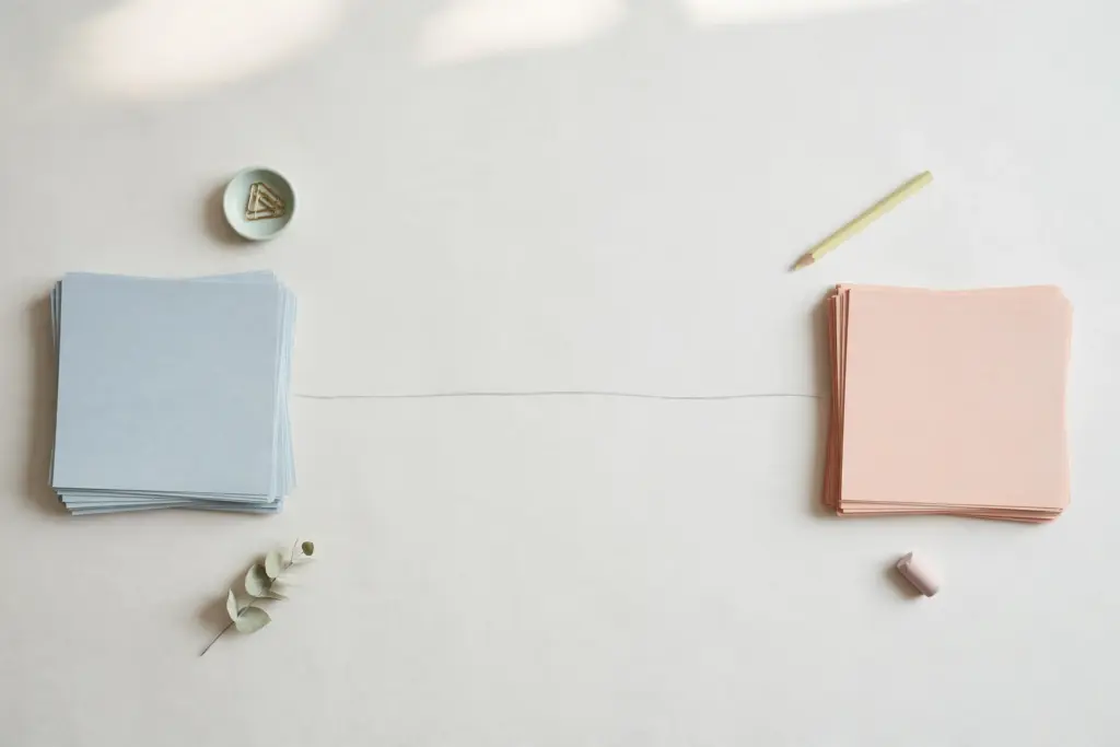 Flat-lay: two stacks of blank paper, pale blue on the left and soft peach on the right, separated by one fine drawn line on a pale off-white desk, with a pencil and a small dish of paperclips nearby