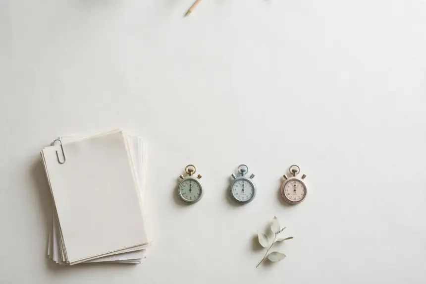 Flat-lay: a neat stack of blank paper reports beside a row of three small pastel stopwatches in mint, blue and pink on a pale off-white background