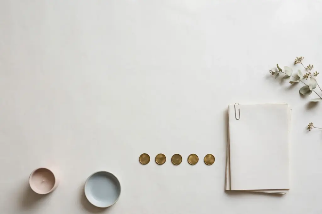 Flat-lay: five small brass counters lined up in a row next to a stack of blank paper reports on a pale off-white desk, with two small pastel dishes and a dried flower sprig nearby