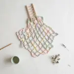 Flat-lay: a safety net woven from pastel ribbons in pink, mint, yellow and blue spread beneath a small wooden ladder on a pale off-white background, with a pencil and a small green bowl at the edges