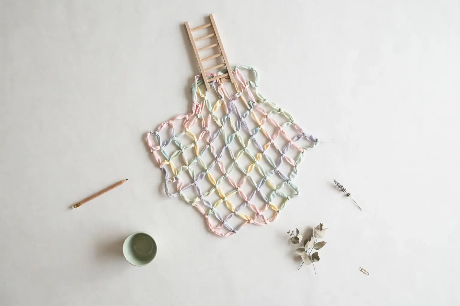 Flat-lay: a safety net woven from pastel ribbons in pink, mint, yellow and blue spread beneath a small wooden ladder on a pale off-white background, with a pencil and a small green bowl at the edges