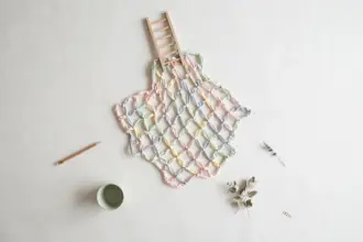 Flat-lay: a safety net woven from pastel ribbons in pink, mint, yellow and blue spread beneath a small wooden ladder on a pale off-white background, with a pencil and a small green bowl at the edges