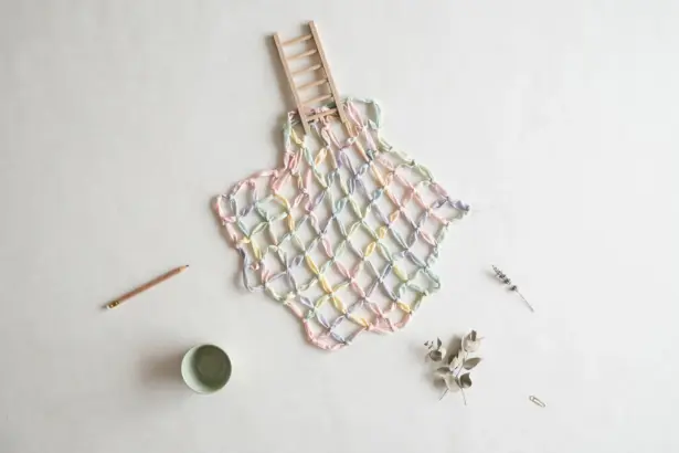 Flat-lay: a safety net woven from pastel ribbons in pink, mint, yellow and blue spread beneath a small wooden ladder on a pale off-white background, with a pencil and a small green bowl at the edges