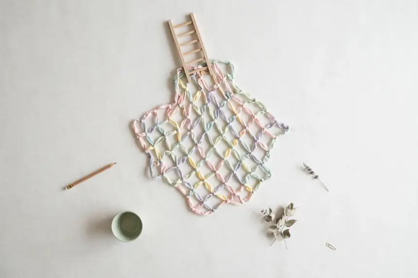 Flat-lay: a safety net woven from pastel ribbons in pink, mint, yellow and blue spread beneath a small wooden ladder on a pale off-white background, with a pencil and a small green bowl at the edges