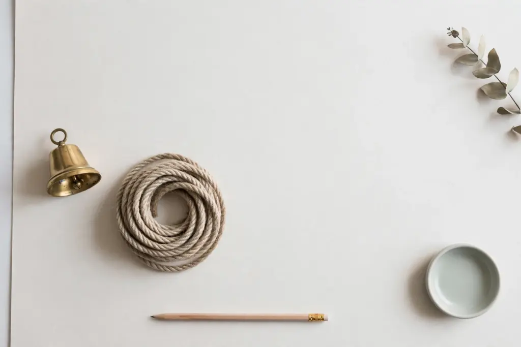 Flat-lay: a small brass bell beside a neatly coiled rope on a pale off-white desk, with a long pencil below and a small pale green dish in the corner