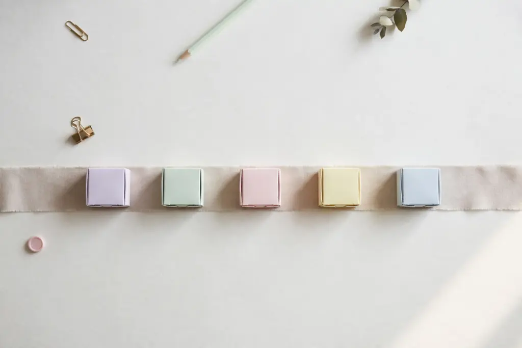 Flat-lay: five small pastel boxes in lavender, mint, pink, yellow and blue waiting in a neat row on a pale fabric conveyor ribbon across an off-white desk