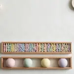 Flat-lay: two long wooden trays side by side on an off-white background, the top one filled with many tiny pastel beads and the lower one holding four large pastel balls