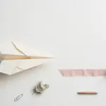 Flat-lay: a paper airplane on a pale off-white desk pointing toward a pink ribbon runway that ends at a small brass bell, with a paperclip and eucalyptus leaf nearby
