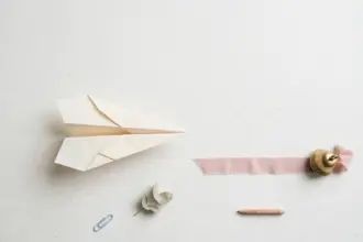 Flat-lay: a paper airplane on a pale off-white desk pointing toward a pink ribbon runway that ends at a small brass bell, with a paperclip and eucalyptus leaf nearby