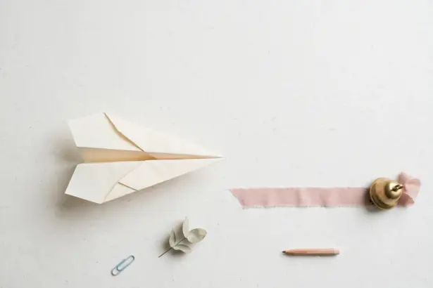 Flat-lay: a paper airplane on a pale off-white desk pointing toward a pink ribbon runway that ends at a small brass bell, with a paperclip and eucalyptus leaf nearby