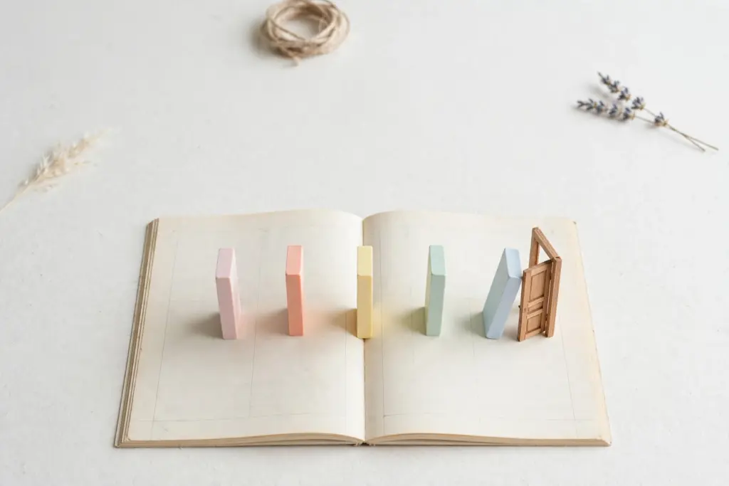 Flat-lay: five pastel dominoes in pink, peach, yellow, mint and blue standing in a row across an open blank ledger, the last one nudging a tiny wooden door ajar