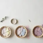 Flat-lay: three small woven baskets on an off-white background holding blank paper tags sorted from pale cream and pink to deeper lavender shades