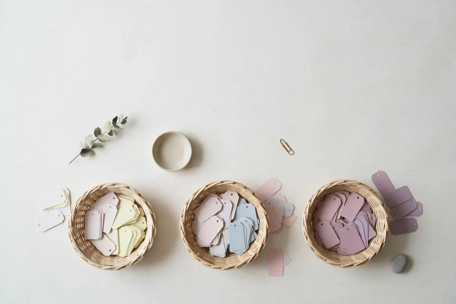 Flat-lay: three small woven baskets on an off-white background holding blank paper tags sorted from pale cream and pink to deeper lavender shades