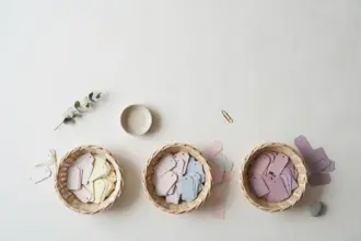 Flat-lay: three small woven baskets on an off-white background holding blank paper tags sorted from pale cream and pink to deeper lavender shades