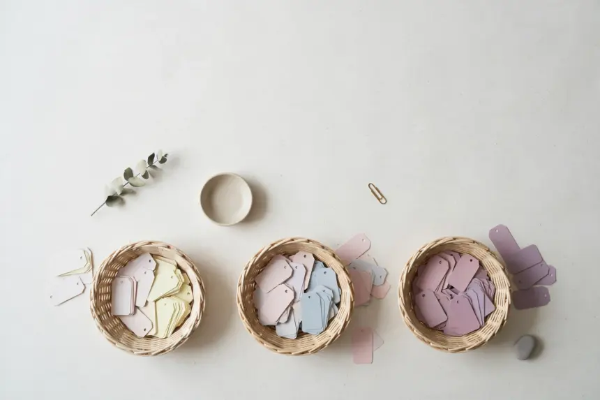 Flat-lay: three small woven baskets on an off-white background holding blank paper tags sorted from pale cream and pink to deeper lavender shades
