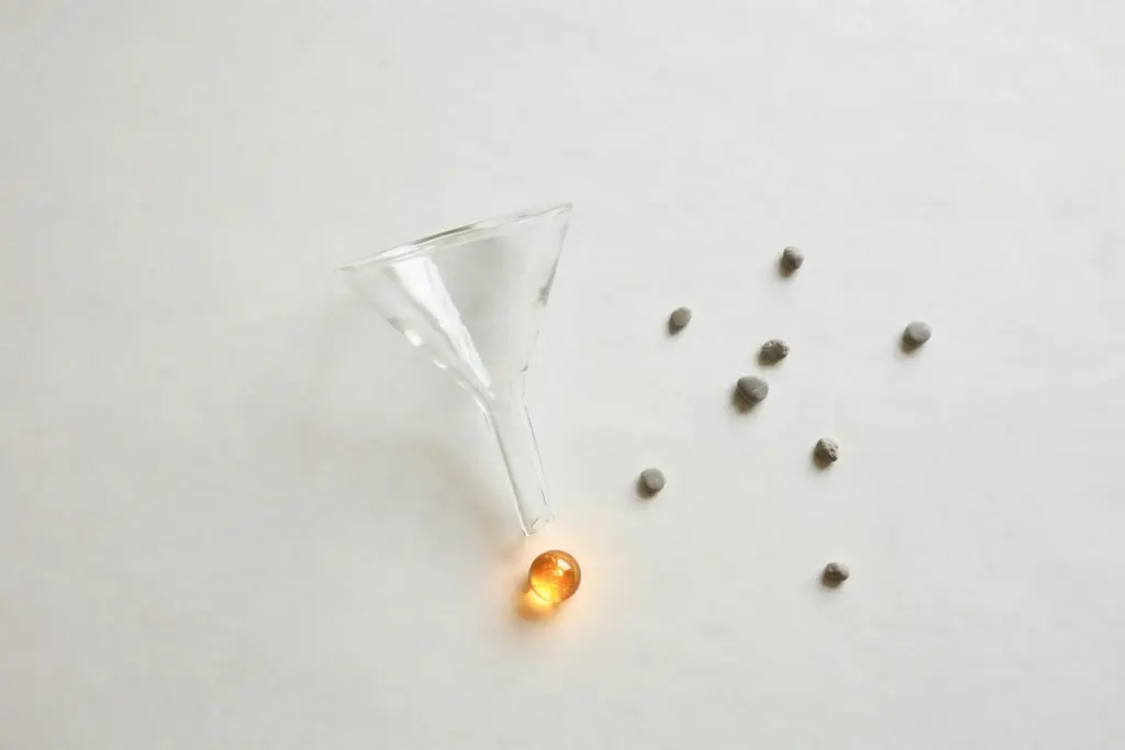 Flat-lay: a clear glass funnel resting above a single glowing amber marble on white paper, with dull grey pebbles scattered set aside on the right