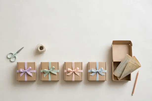 Flat-lay: a row of four small kraft boxes each tied with a different pastel ribbon beside an open fifth box with folded paper plans peeking out, with scissors and twine nearby