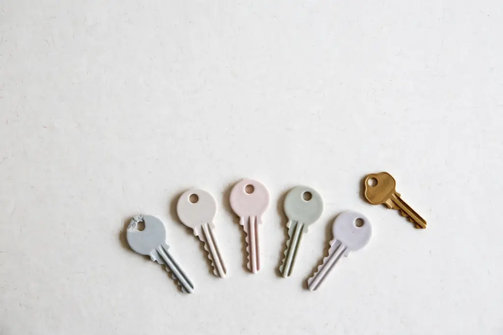 Flat-lay: one brass key resting beside five pale pastel ceramic key copies fanned out in a gentle arc on an off-white background, one copy slightly chipped