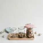 Flat-lay: three clear glass jars of coins on a wooden tray — one nearly empty, one half full and one full to the brim with a small pink paper umbrella resting on top