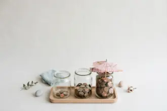 Flat-lay: three clear glass jars of coins on a wooden tray — one nearly empty, one half full and one full to the brim with a small pink paper umbrella resting on top