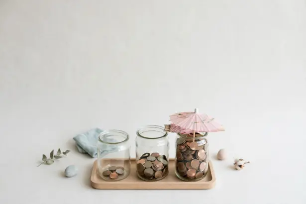 Flat-lay: three clear glass jars of coins on a wooden tray — one nearly empty, one half full and one full to the brim with a small pink paper umbrella resting on top