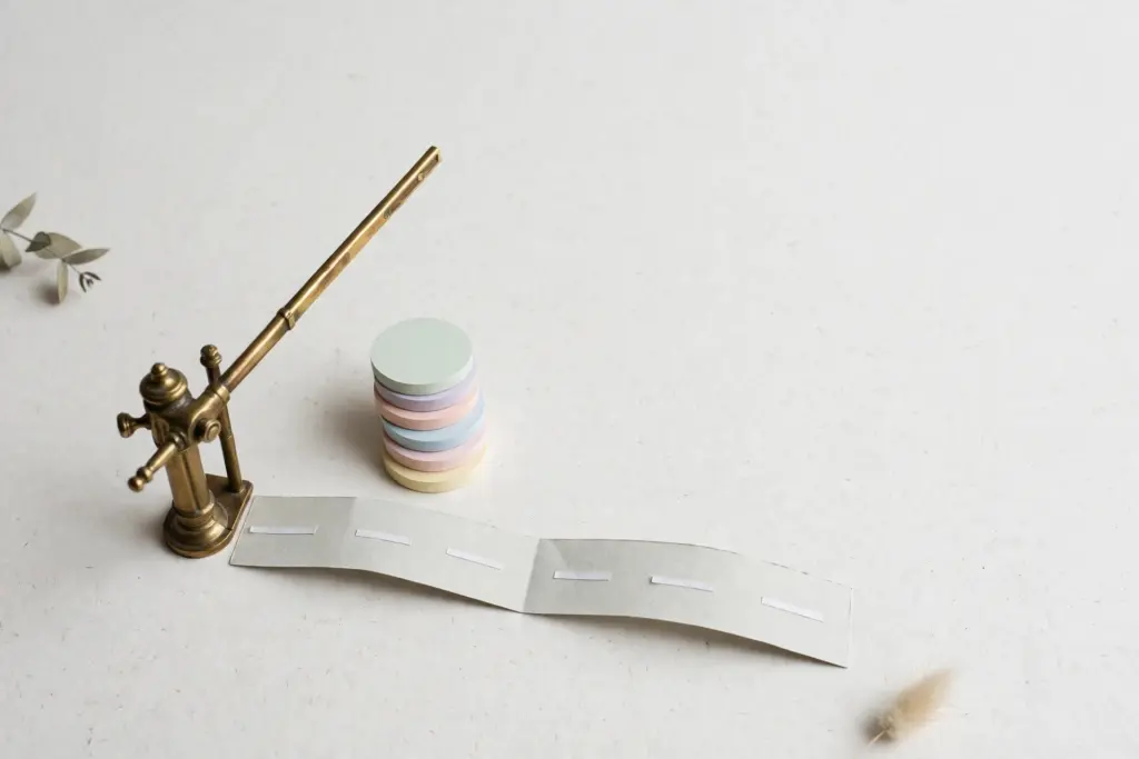 Flat-lay: a vintage brass toll gate with its arm lifted over a small pale paper road, a neat stack of round pastel toll tokens beside the gate post