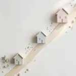 Flat-lay: three small model houses of increasing size in sage, blue-grey and blush arranged along a rising beige ribbon street on an off-white background