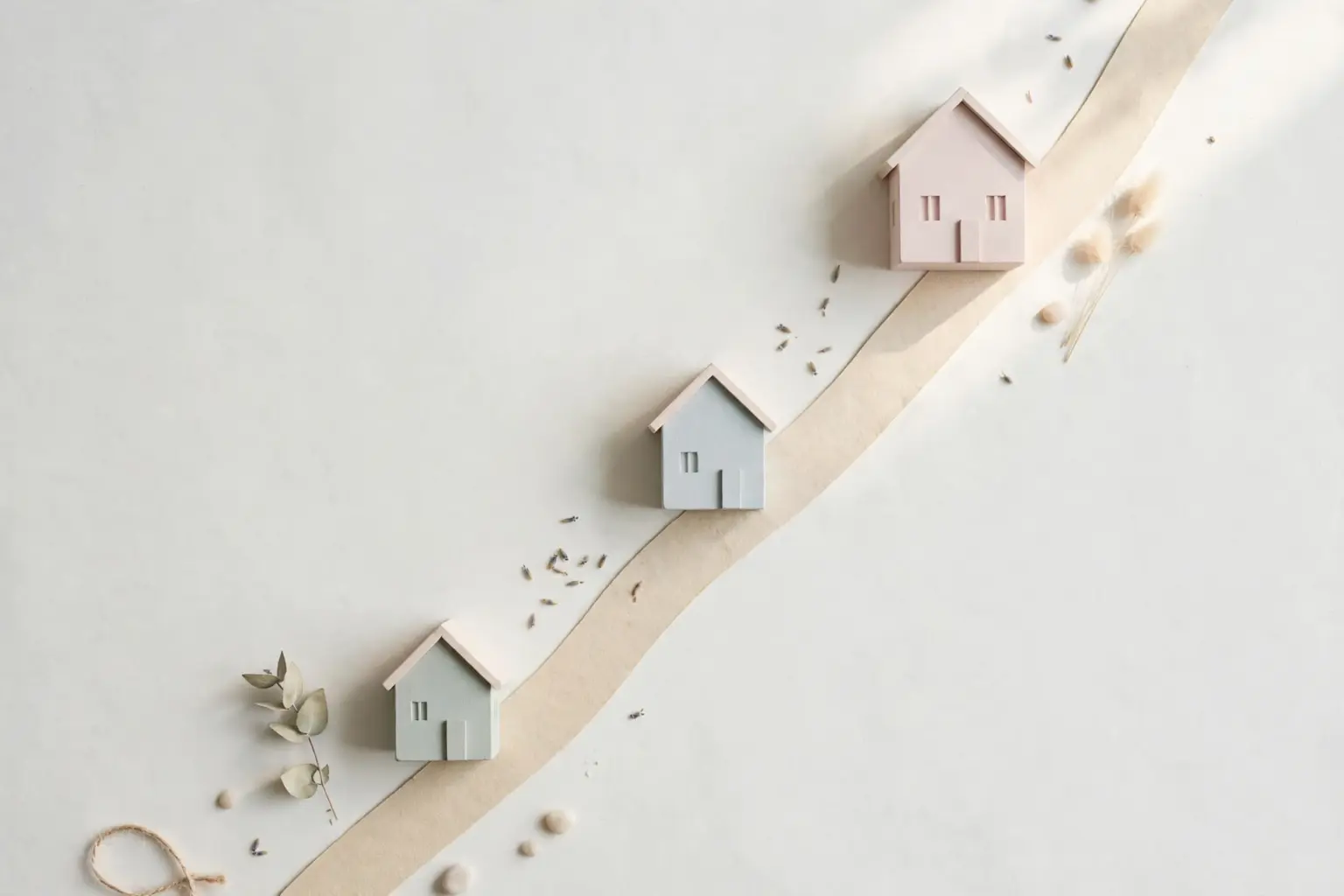 Flat-lay: three small model houses of increasing size in sage, blue-grey and blush arranged along a rising beige ribbon street on an off-white background