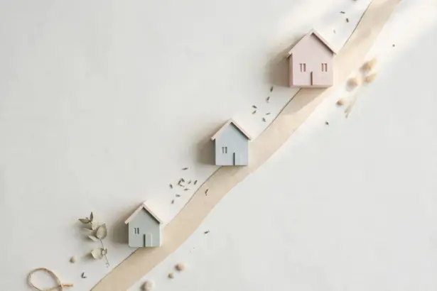 Flat-lay: three small model houses of increasing size in sage, blue-grey and blush arranged along a rising beige ribbon street on an off-white background