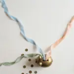 Flat-lay: three thin pastel ribbons in blue, peach and sage streaming across an off-white background and converging into a small round brass piggy bank, with scattered coins and a green leaf at the edge