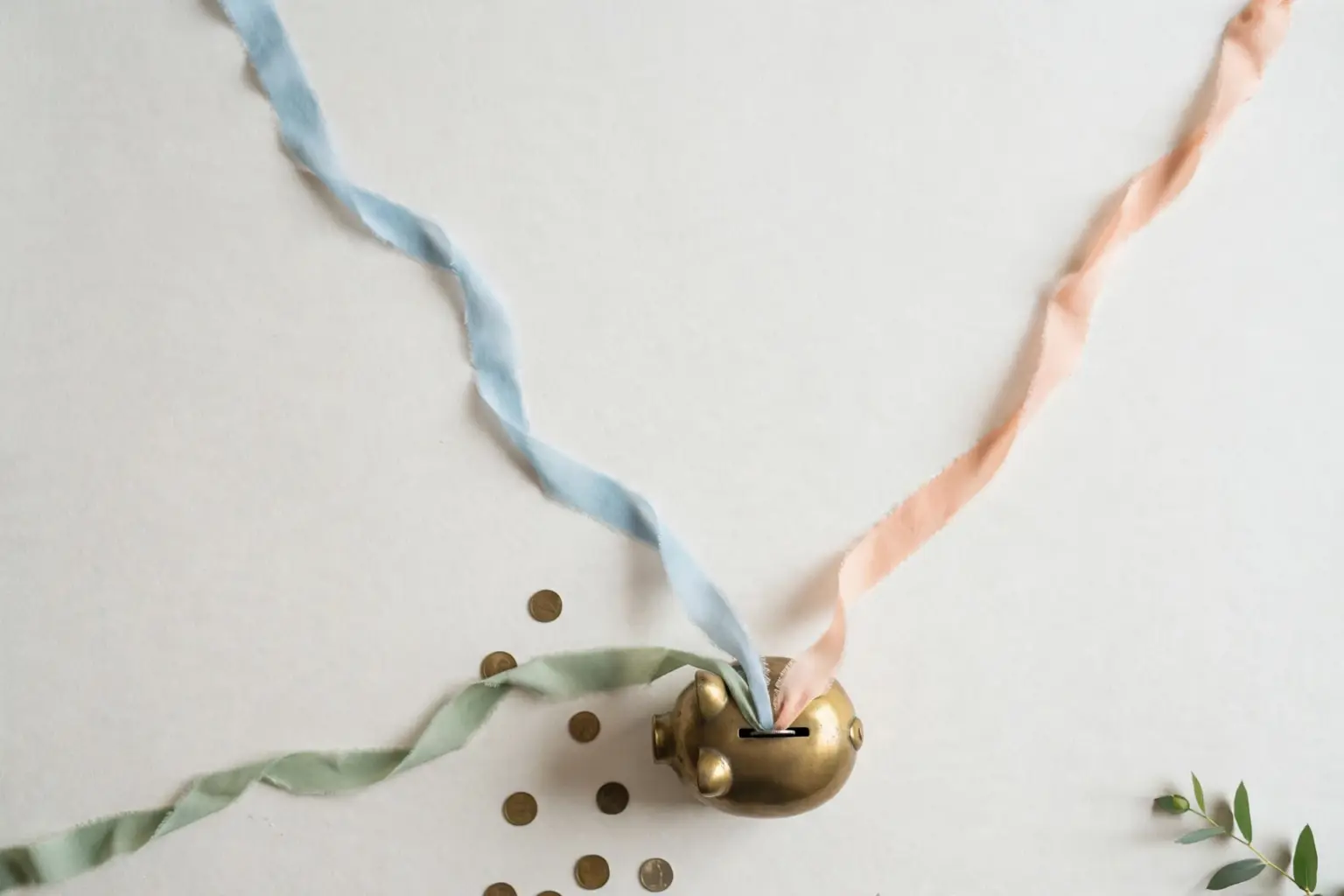 Flat-lay: three thin pastel ribbons in blue, peach and sage streaming across an off-white background and converging into a small round brass piggy bank, with scattered coins and a green leaf at the edge