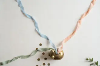 Flat-lay: three thin pastel ribbons in blue, peach and sage streaming across an off-white background and converging into a small round brass piggy bank, with scattered coins and a green leaf at the edge